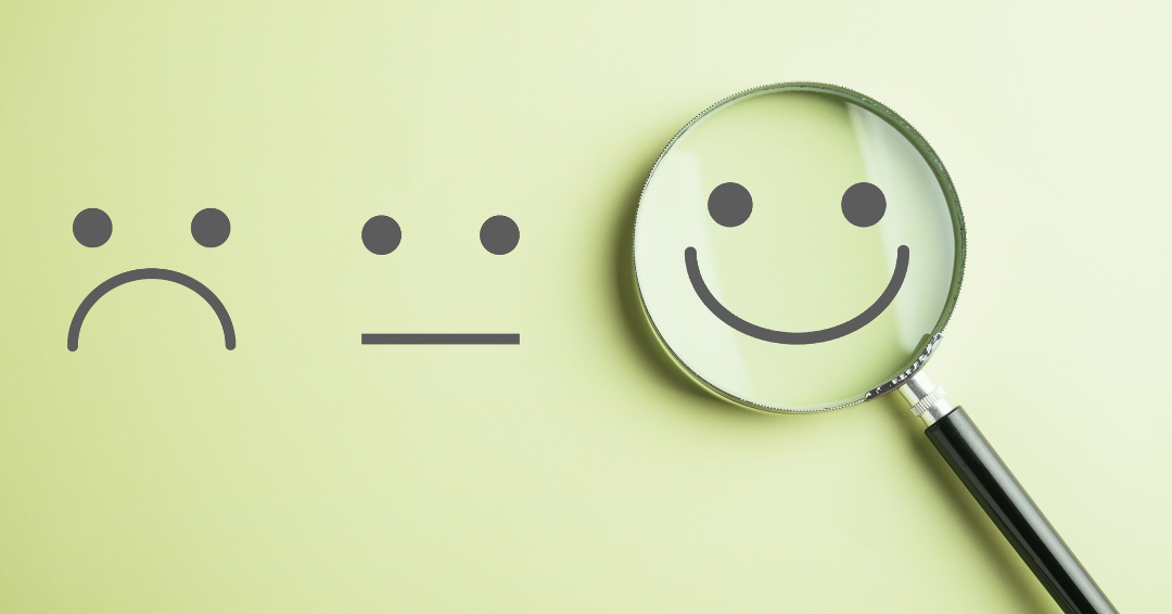 Photo of three smiley faces on yellow paper - one said, one neutral, one happy. With the happy face highlighted by a magnifying glass.