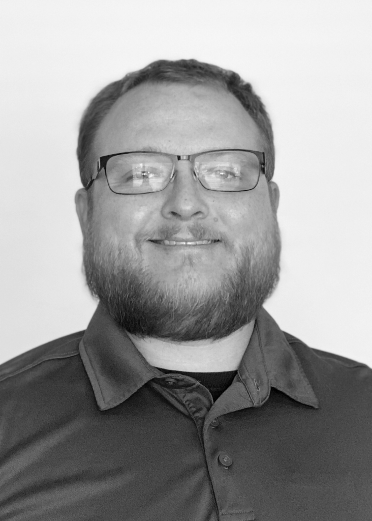 Headshot for Charles Wiltgen- a white man in his 30s with a beard, mustache, and black rectangular glass. 