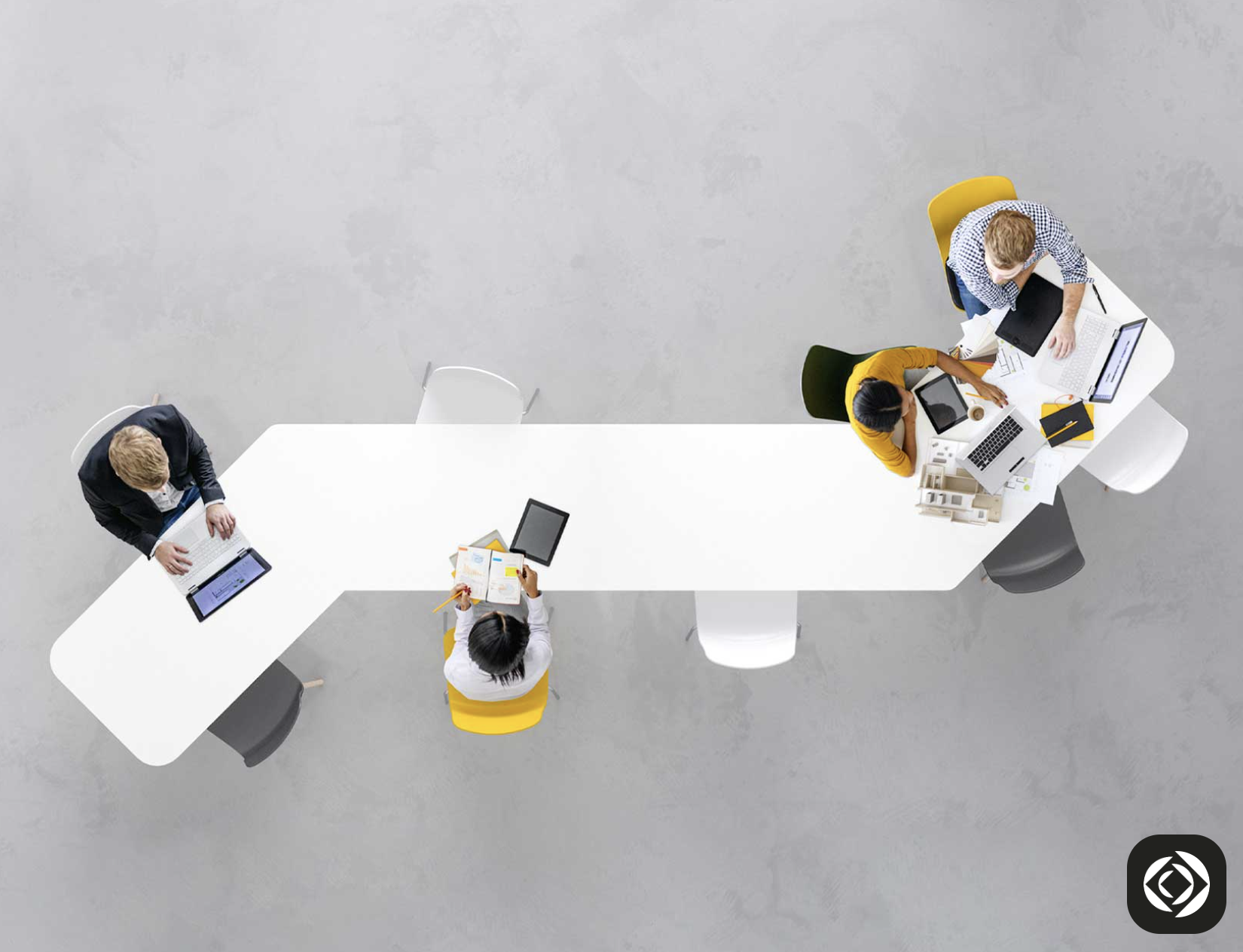 An overhead photograph of four coworkers using computers at a white, elongated table. It's rectangular but angles at each end, in opposite directions. The chairs are a mix of white, yellow, and dark gray.