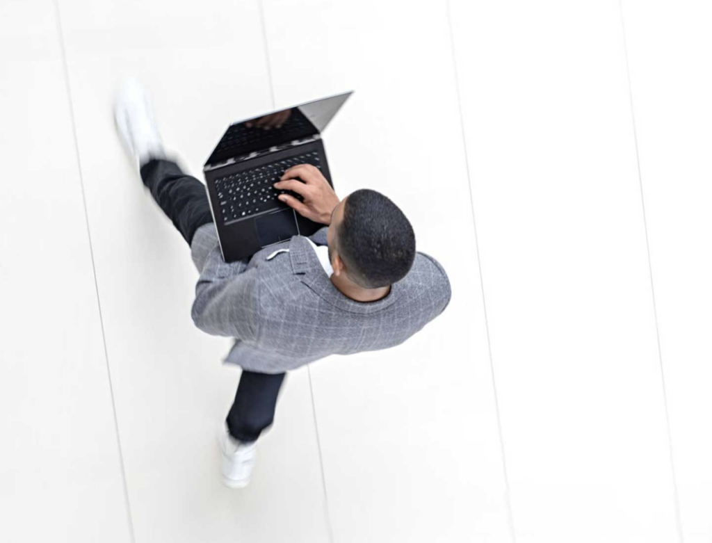 An overhead photograph of a man walking while using his laptop computer. He's wearing a gray jacket with a white grid. He's walking quickly because his sneakers are blurred.