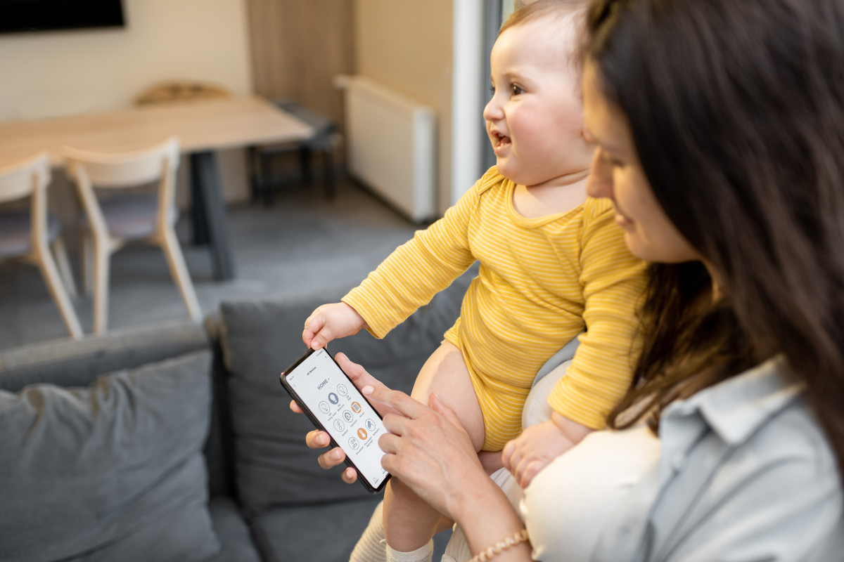 Photo of mother holding a baby on her hip. The baby is wearing a yellow long-sleeved onesie. The mother is using her mobile phone to review details from the baby's daycare center.