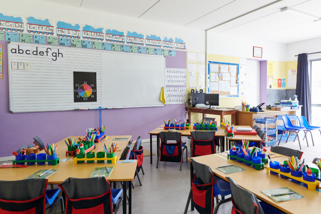 Photo of an empty but pleasantly lit kindergarten classroom. There are several desks each with colorful cups of art supplies. The whiteboard on the wall shows the letters a, b, c, d, e, f, g, and h. The teacher's desk is in the back left corner of the room. This image helps to illustrate our FileMaker in education article.