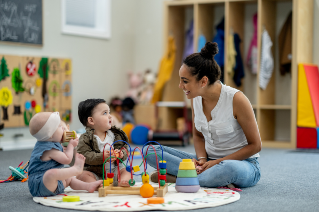 Photo of a daycare center where a teacher and two babies are sitting on a circular rug. The babies are holding toys, looking at the teacher, who is smiling at them. This image helps to illustrate our FileMaker in education article.