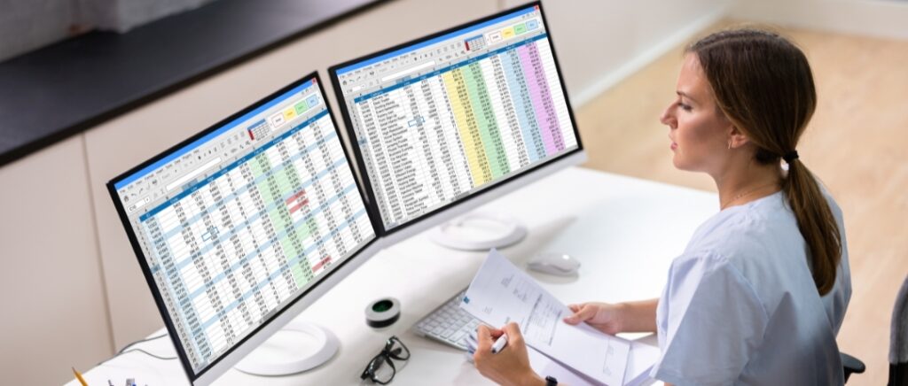 Businesswoman, with hair pulled back in a ponytail, sitting at a white desk, in front of two large computer monitors. Each monitor is displaying spreadsheets with lots of data, with some colorful columns.