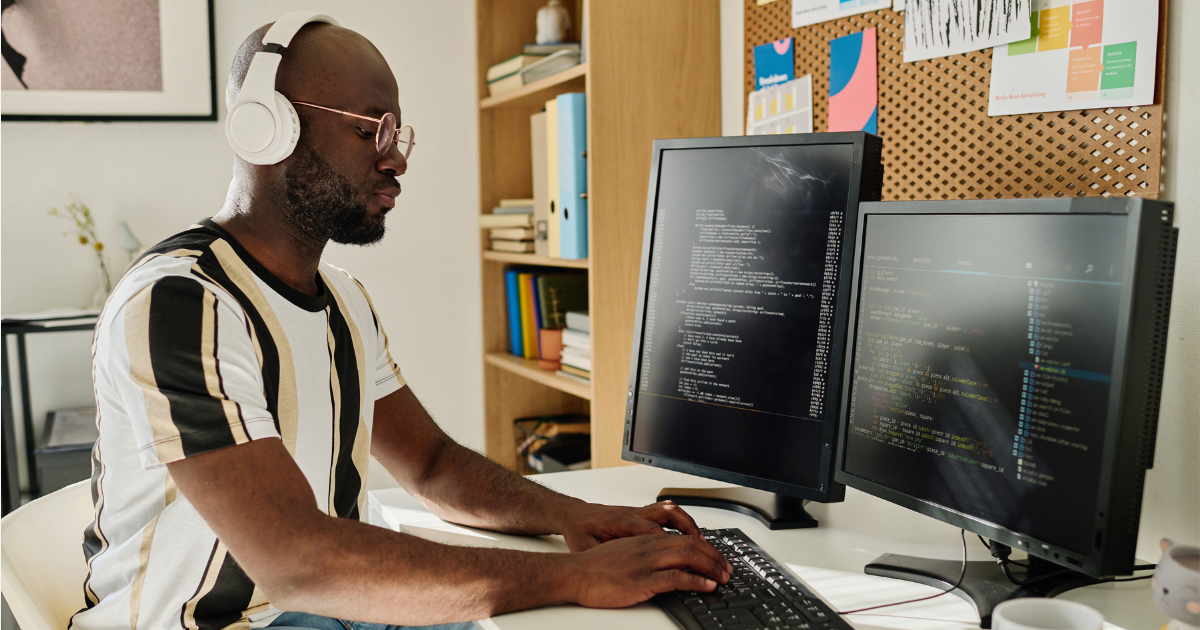 A man sitting at his desk typing on a desktop computer and wearing headphones.